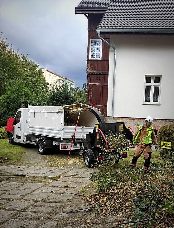 Arborysta w kasku i odzieży ochronnej wkłada gałęzie do rębak-rozdrabniarki, obok stoi biały samochód dostawczy z otwartą paką, na tle budynku mieszkalnego i pochmurnego nieba.